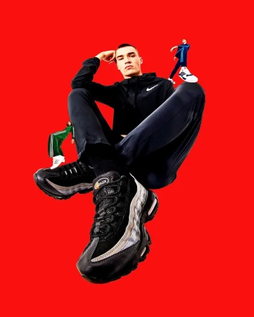 A low-angle shot captures a young man in sportswear seated against a red backdrop, highlighting his black and gray Nike Air Max sneakers.