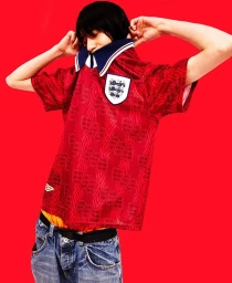 Model wearing a red football jersey and blue jeans posing against a vibrant red background in London by Hanina