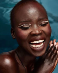 Close-up portrait of a smiling Black woman with jeweled eye makeup and glowing skin against blue water background