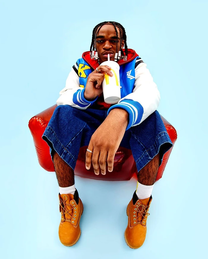 Black man with braided hair wearing a blue varsity jacket and denim jeans seated in a red inflatable chair holding a drink