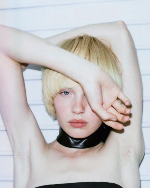 Close-up studio portrait of an androgynous young adult with blond blunt-cut hair wearing black tube top and wide choker with white background.