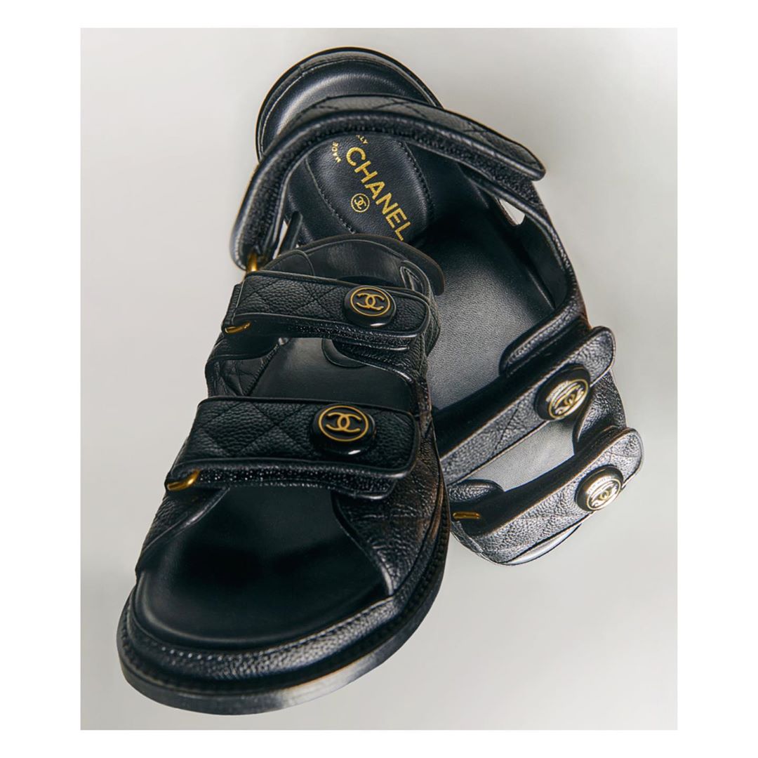 Pair of black leather Chanel sandals with quilted straps, gold-tone hardware, and adjustable velcro closures, displayed on a white surface.