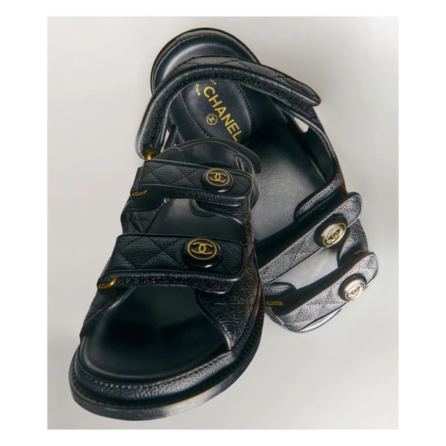 Pair of black leather Chanel sandals with quilted straps, gold-tone hardware, and adjustable velcro closures, displayed on a white surface.
