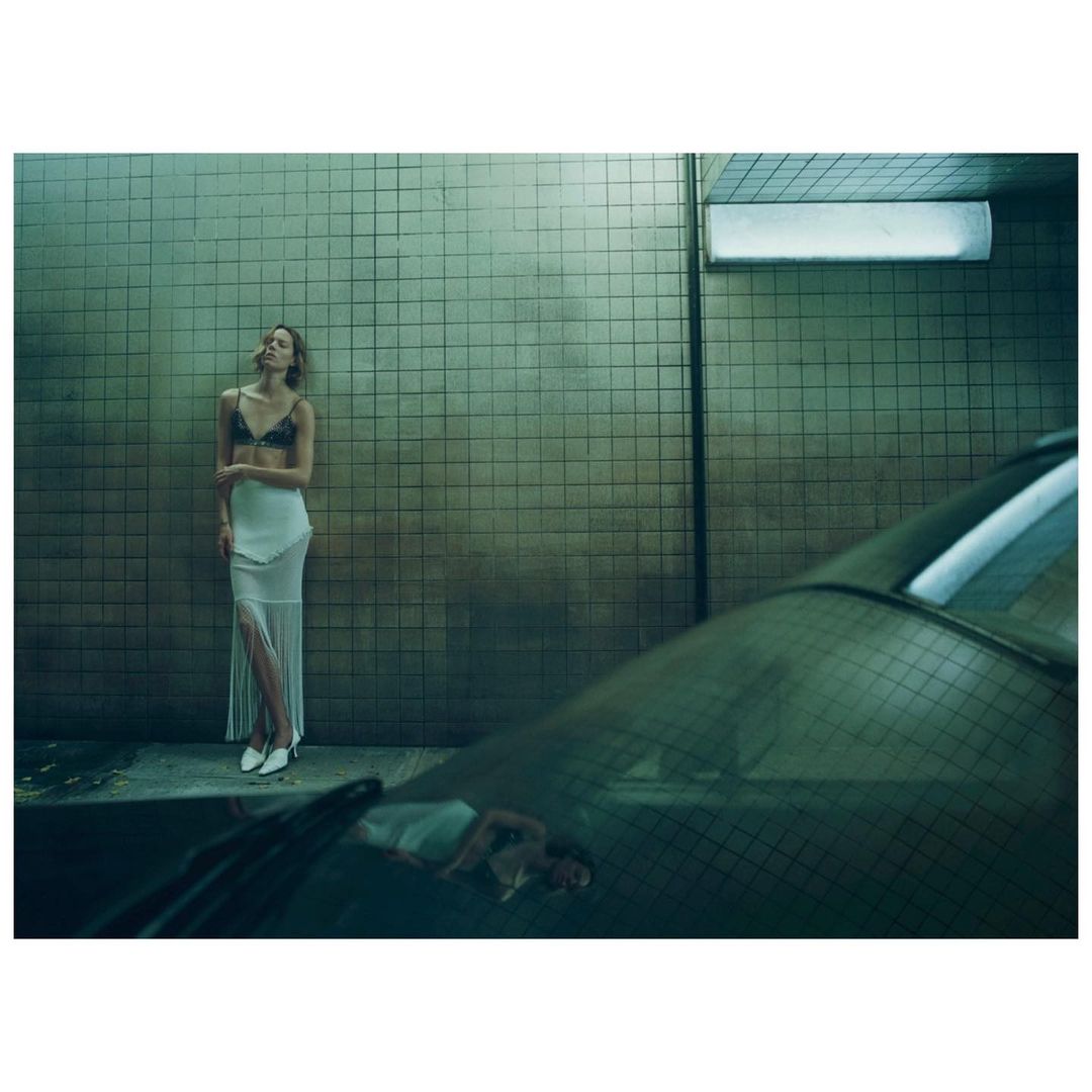 A woman in a lingerie top and skirt stands against a tiled wall with a car bumper in the foreground, styled fashion photograph.