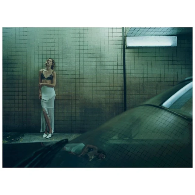 A woman in a lingerie top and skirt stands against a tiled wall with a car bumper in the foreground, styled fashion photograph.