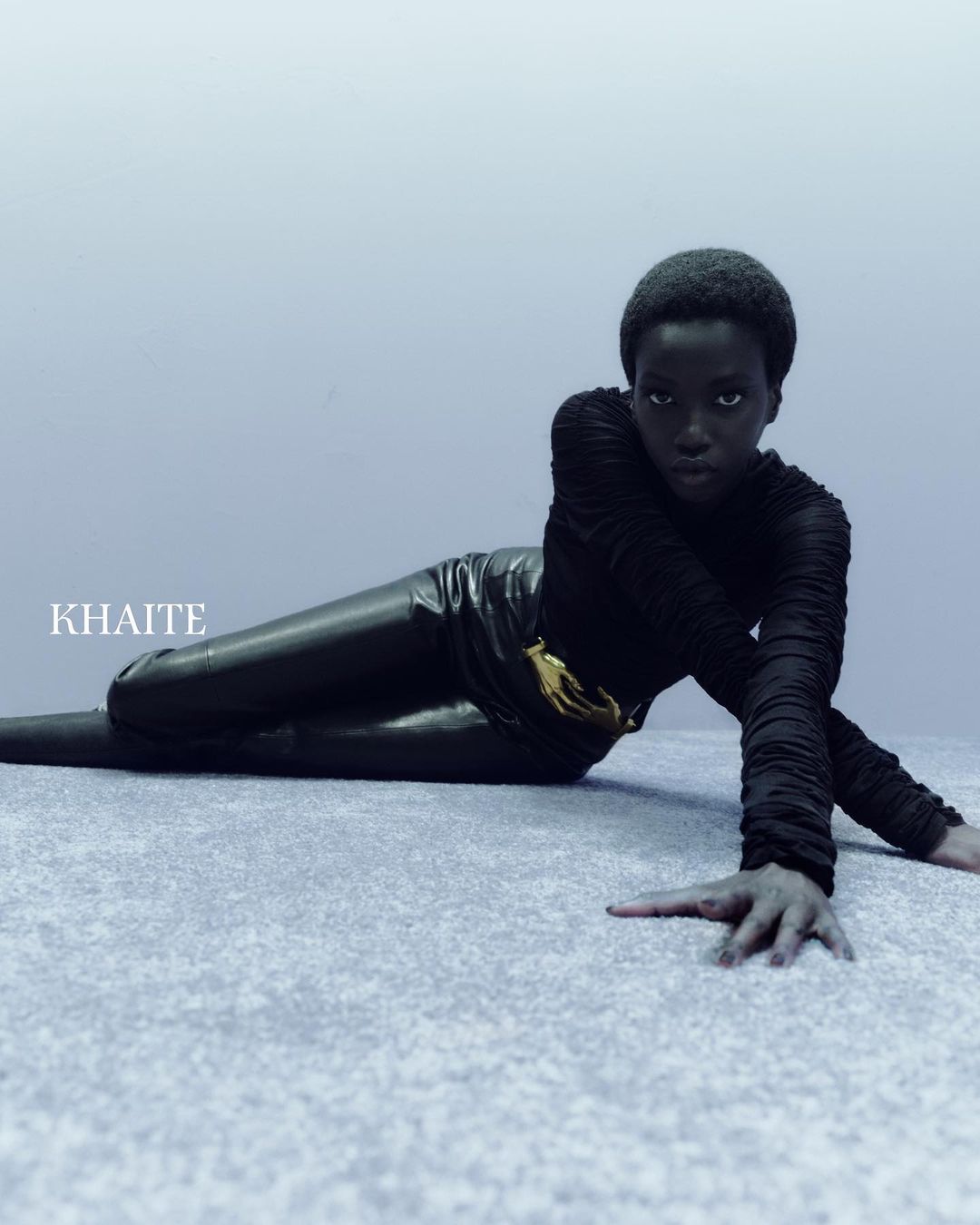 A Black woman is lying down in a black long-sleeve and black leggings with a gold belt. The model is indoors with studio lighting.