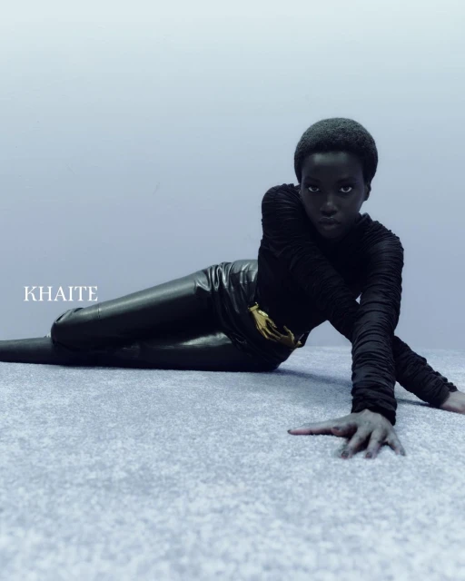 A Black woman is lying down in a black long-sleeve and black leggings with a gold belt. The model is indoors with studio lighting.