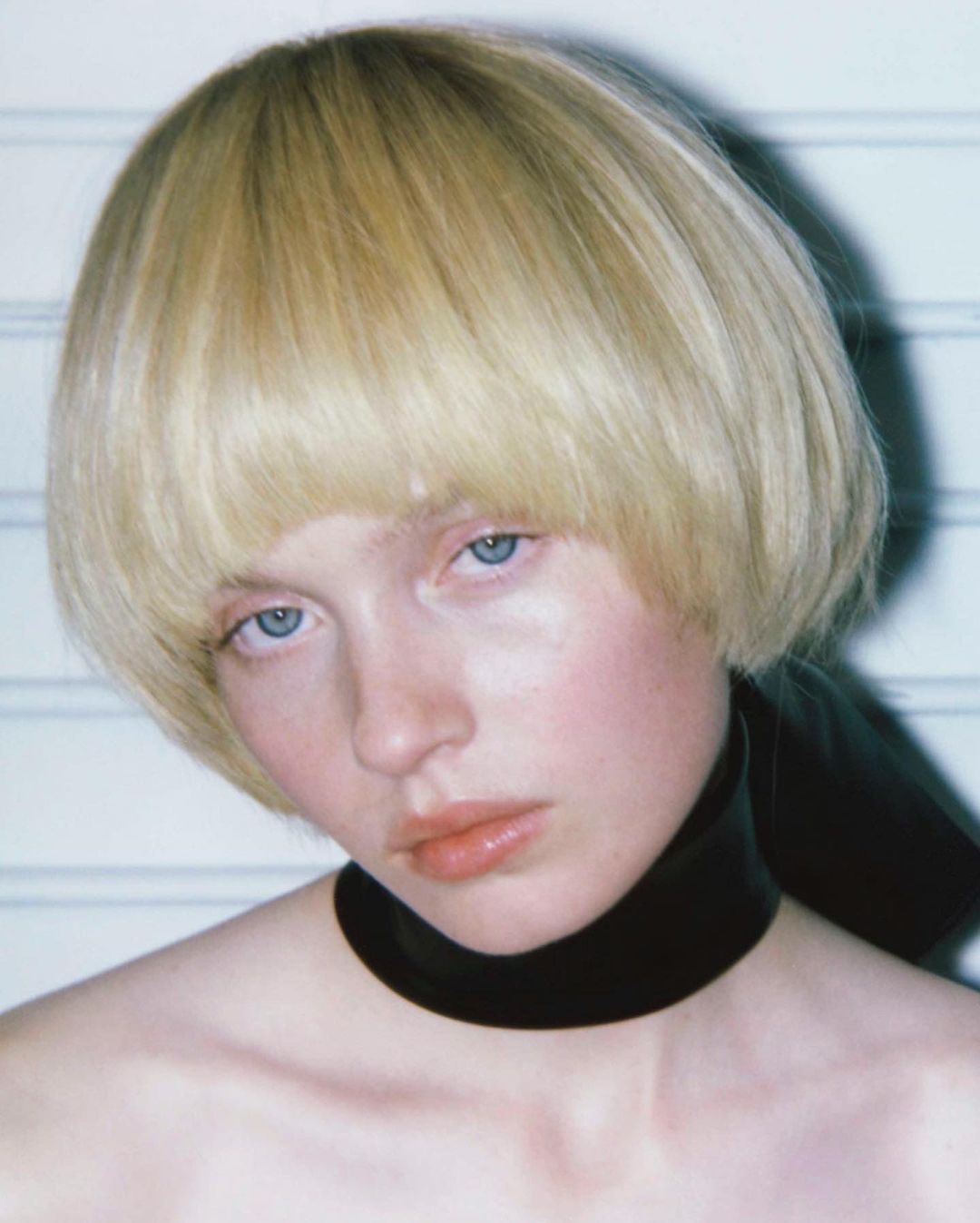Close-up portrait of a young, fair-skinned woman with a blonde bowl cut and light blue eyes wearing a black choker against a white background.