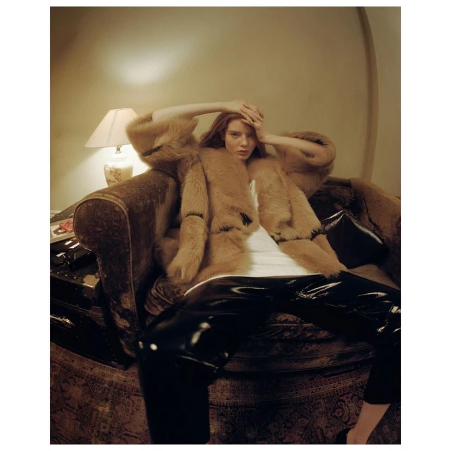 A woman in a fur coat is sitting on an ornate chair with her hands above her head in an indoor setting with warm, soft lighting.