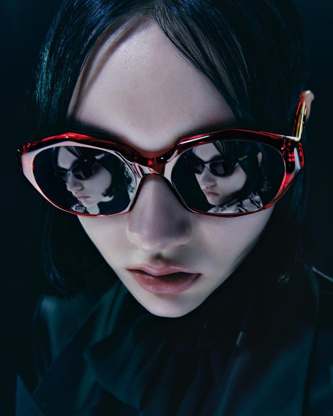 A young adult with short dark hair wearing red sunglasses that reflect another face with sunglasses, close-up shot, dark background.