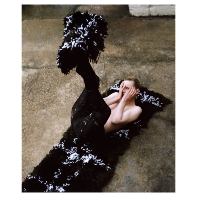 Artistic fashion photo of a shirtless male model lying on a black and white shag rug with feathered headdress and face framing.