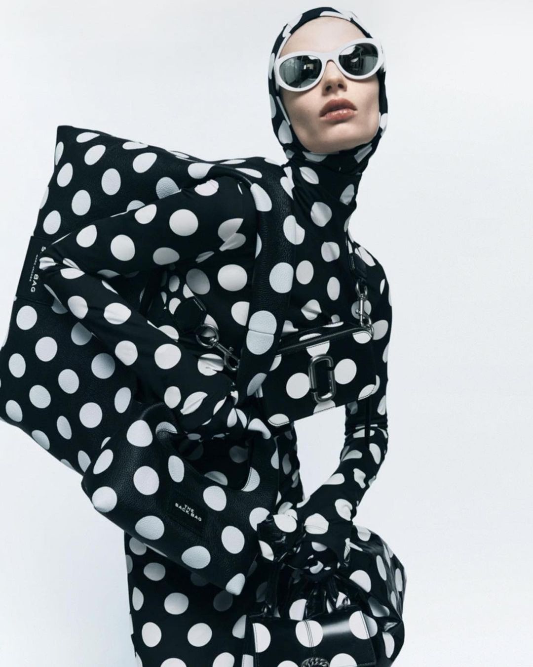 A fashion shot featuring a model in a black and white polka-dot bodysuit, sunglasses, and multiple black purses, against a white backdrop.