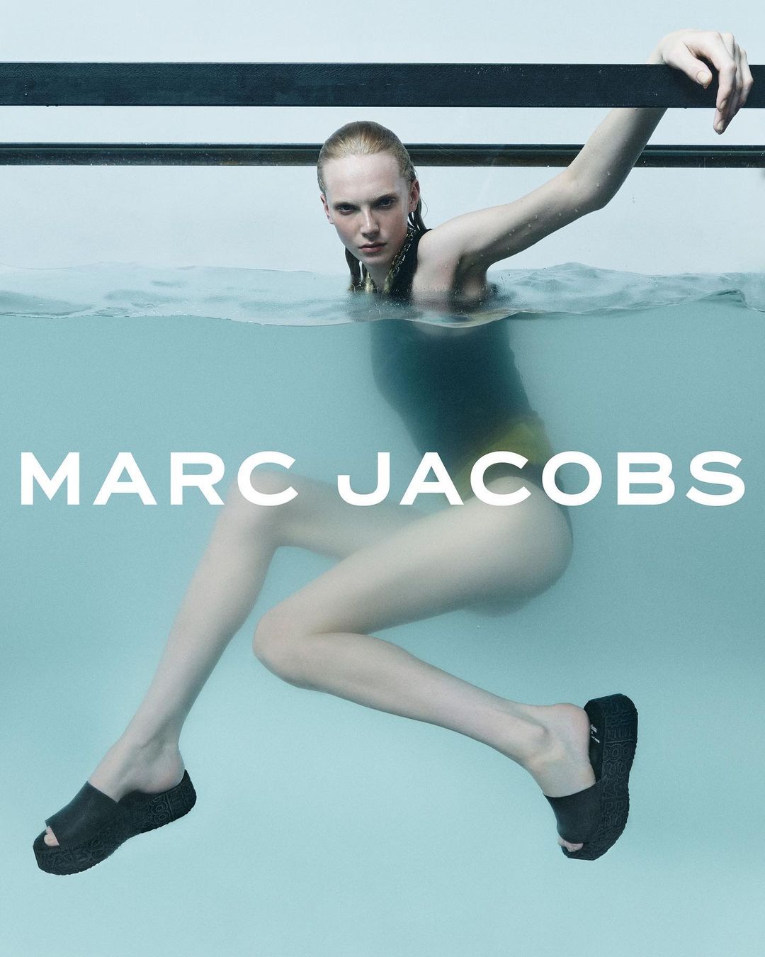 Fashion ad featuring a woman partially submerged in water wearing a black swimsuit and black platform sandals, with visible logo.