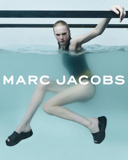 Fashion ad featuring a woman partially submerged in water wearing a black swimsuit and black platform sandals, with visible logo.