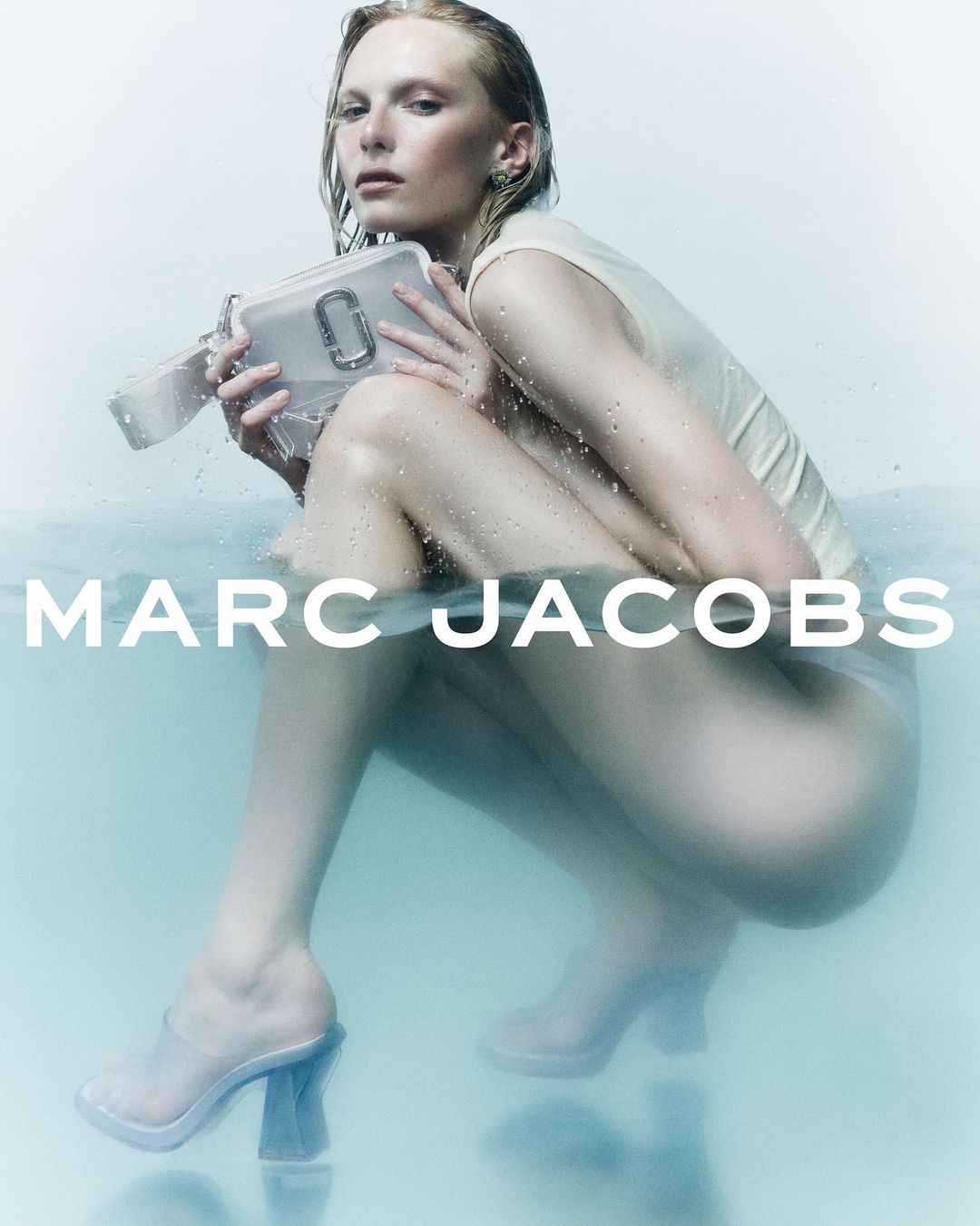 Fashion shot of a blonde model partially submerged in water holding a purse with the Marc Jacobs logo, and wearing clear heeled shoes.