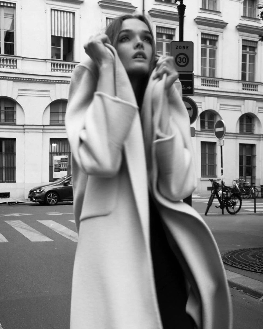 Black and white portrait of an adult woman wearing a coat on a city street with buildings, a car, and street signs in the background.