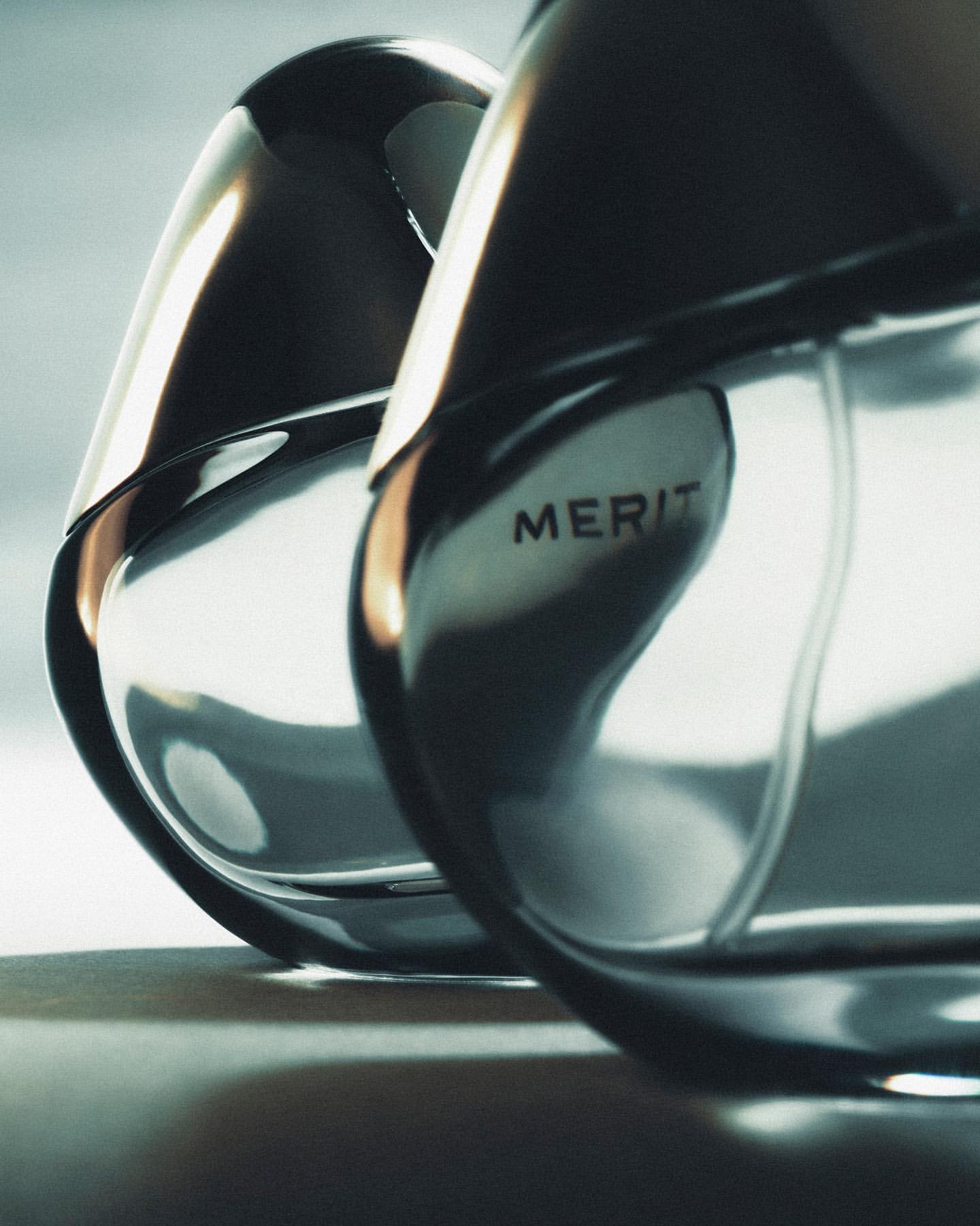 Close-up of two sleek, reflective perfume bottles with black accents and the word 'MERIT' visible, set against a soft gradient backdrop.