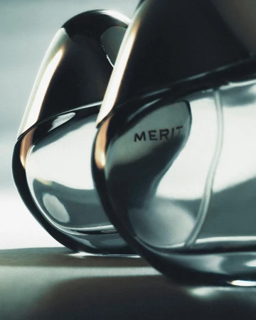 Close-up of two sleek, reflective perfume bottles with black accents and the word 'MERIT' visible, set against a soft gradient backdrop.