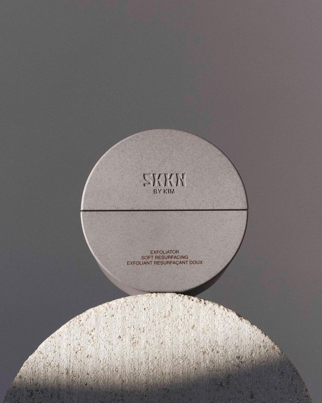 Elevated studio shot featuring a SKKN by Kim exfoliating product atop a textured half-sphere stone against a plain grey backdrop.