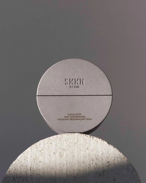 Elevated studio shot featuring a SKKN by Kim exfoliating product atop a textured half-sphere stone against a plain grey backdrop.
