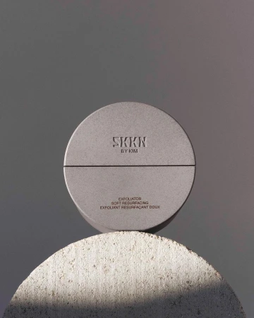 Elevated studio shot featuring a SKKN by Kim exfoliating product atop a textured half-sphere stone against a plain grey backdrop.