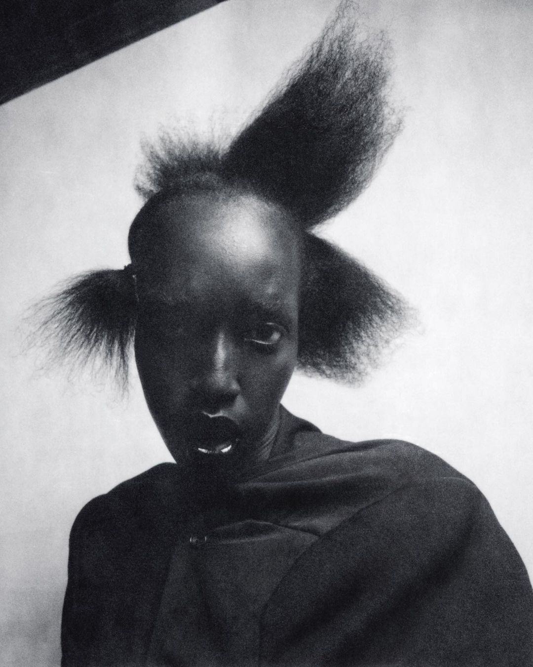 Close-up portrait of a dark-skinned woman with unique hairstyle, wearing a black wrap-style garment, and featuring silver lip and nose piercings.