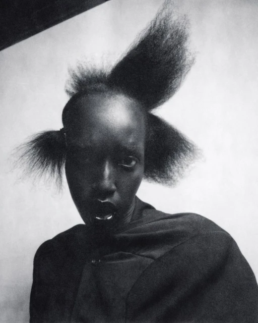 Close-up portrait of a dark-skinned woman with unique hairstyle, wearing a black wrap-style garment, and featuring silver lip and nose piercings.