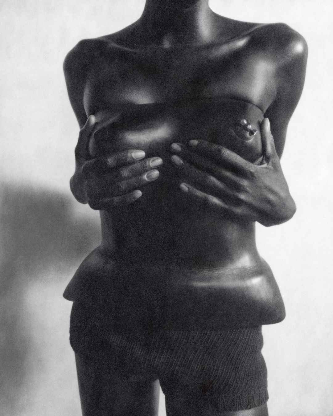 Monochrome studio shot of an African-descent woman covering her breasts with her hands, wearing high-waisted shorts against a white background.