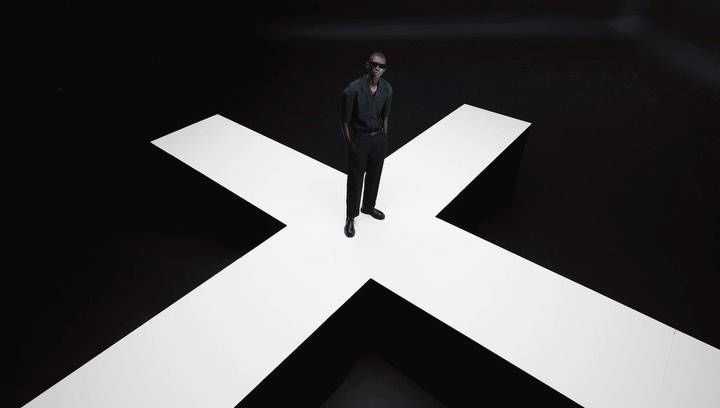 Stylish Black male model in sunglasses poses on a white cross against a black studio backdrop, showcasing contemporary fashion.