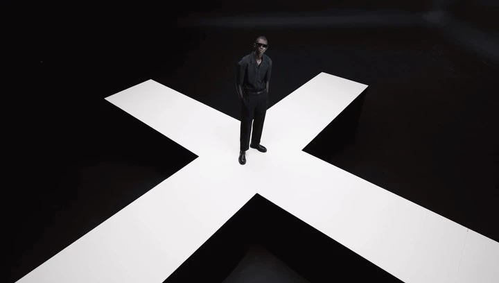 Stylish Black male model in sunglasses poses on a white cross against a black studio backdrop, showcasing contemporary fashion.