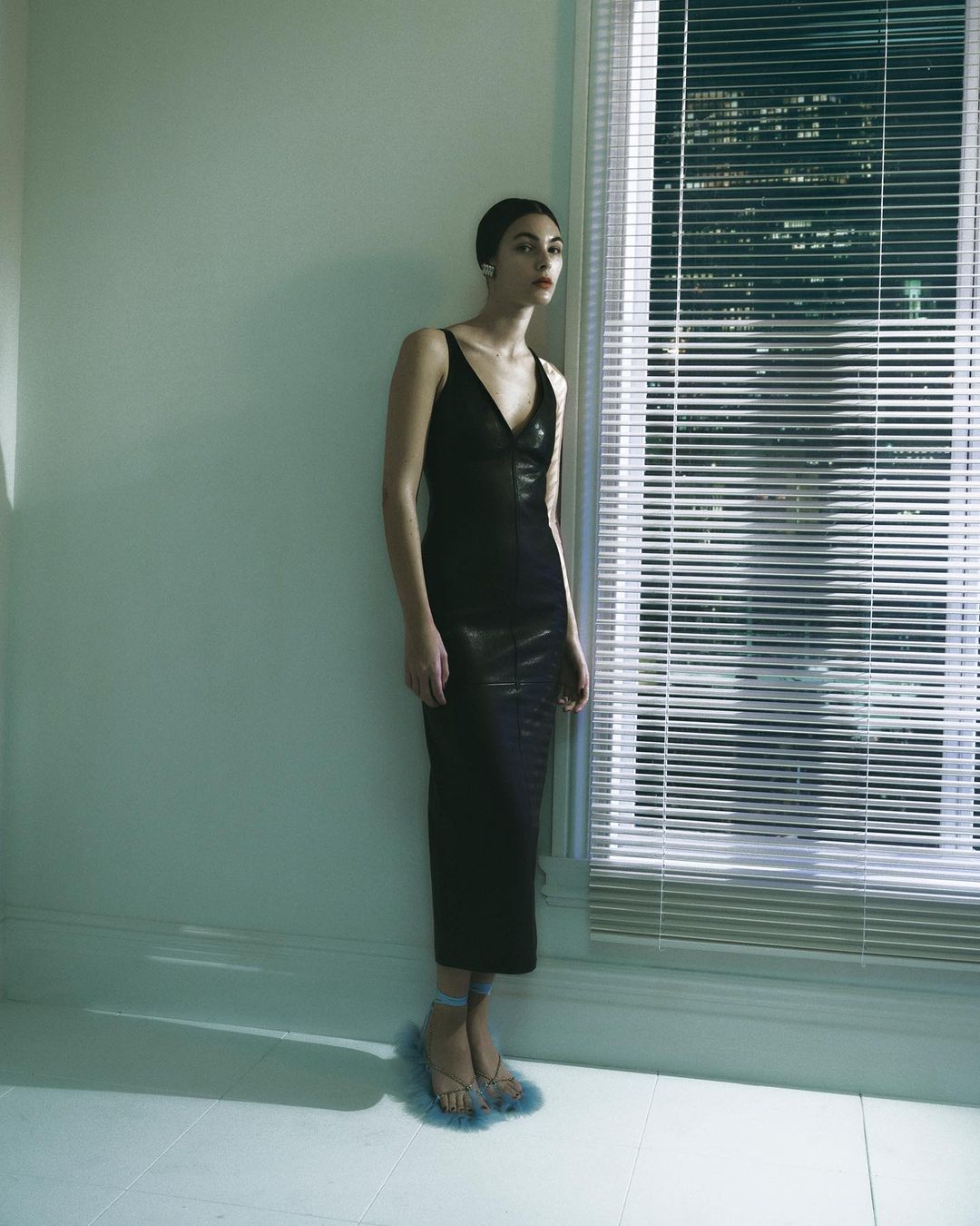 A woman stands near a window in a sleeveless black dress and fur sandals, in a dimly lit interior.