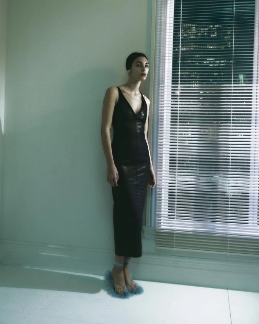 A woman stands near a window in a sleeveless black dress and fur sandals, in a dimly lit interior.