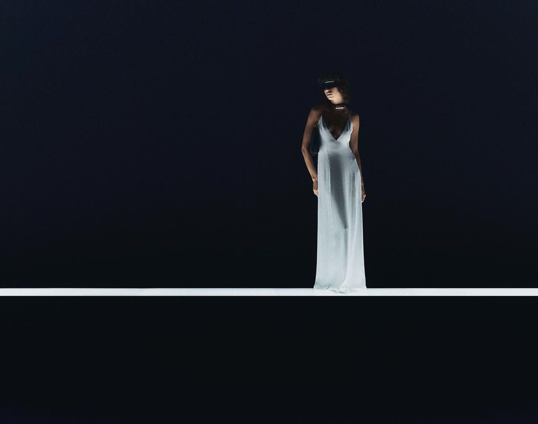 Elegant woman in a white dress stands on a bright white line against a dark background, captured in a studio-lit fashion shot.