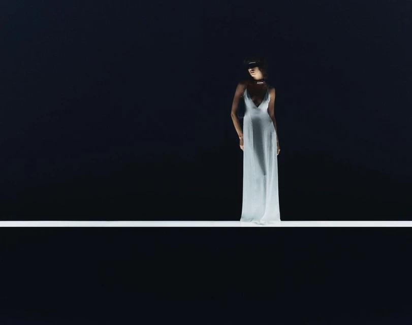 Elegant woman in a white dress stands on a bright white line against a dark background, captured in a studio-lit fashion shot.