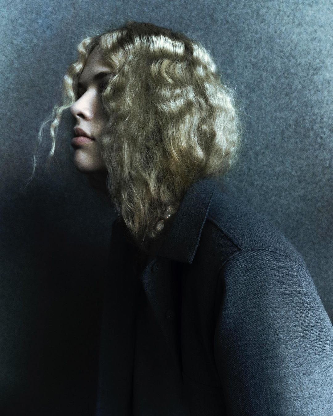 Profile view of an adult female with blonde wavy hair wearing a dark grey collared top, against a textured grey-blue background.