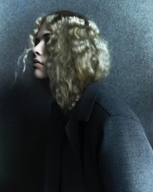Profile view of an adult female with blonde wavy hair wearing a dark grey collared top, against a textured grey-blue background.