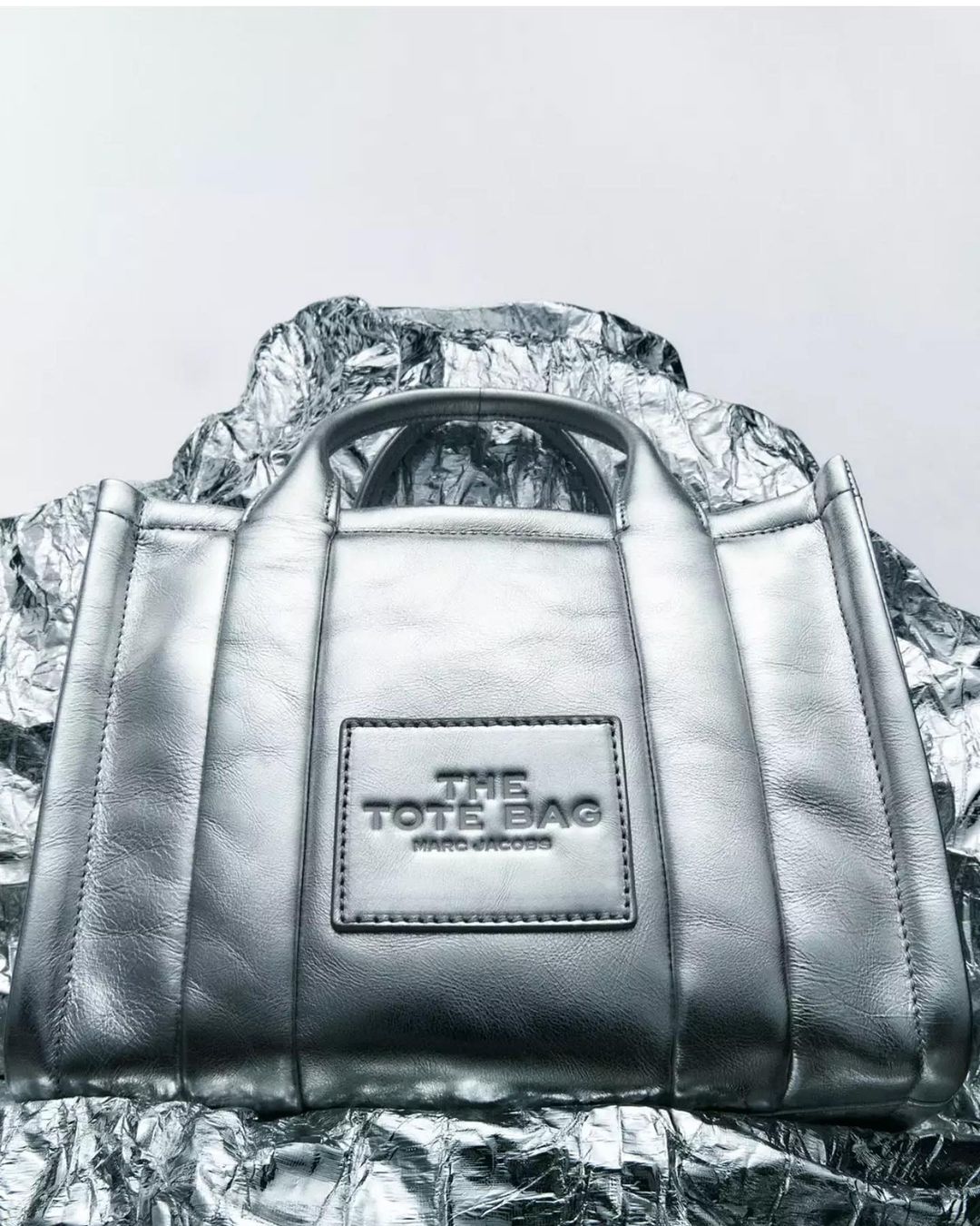Silver metallic tote bag on a crumpled metallic surface, against a plain grey background, product studio photography.