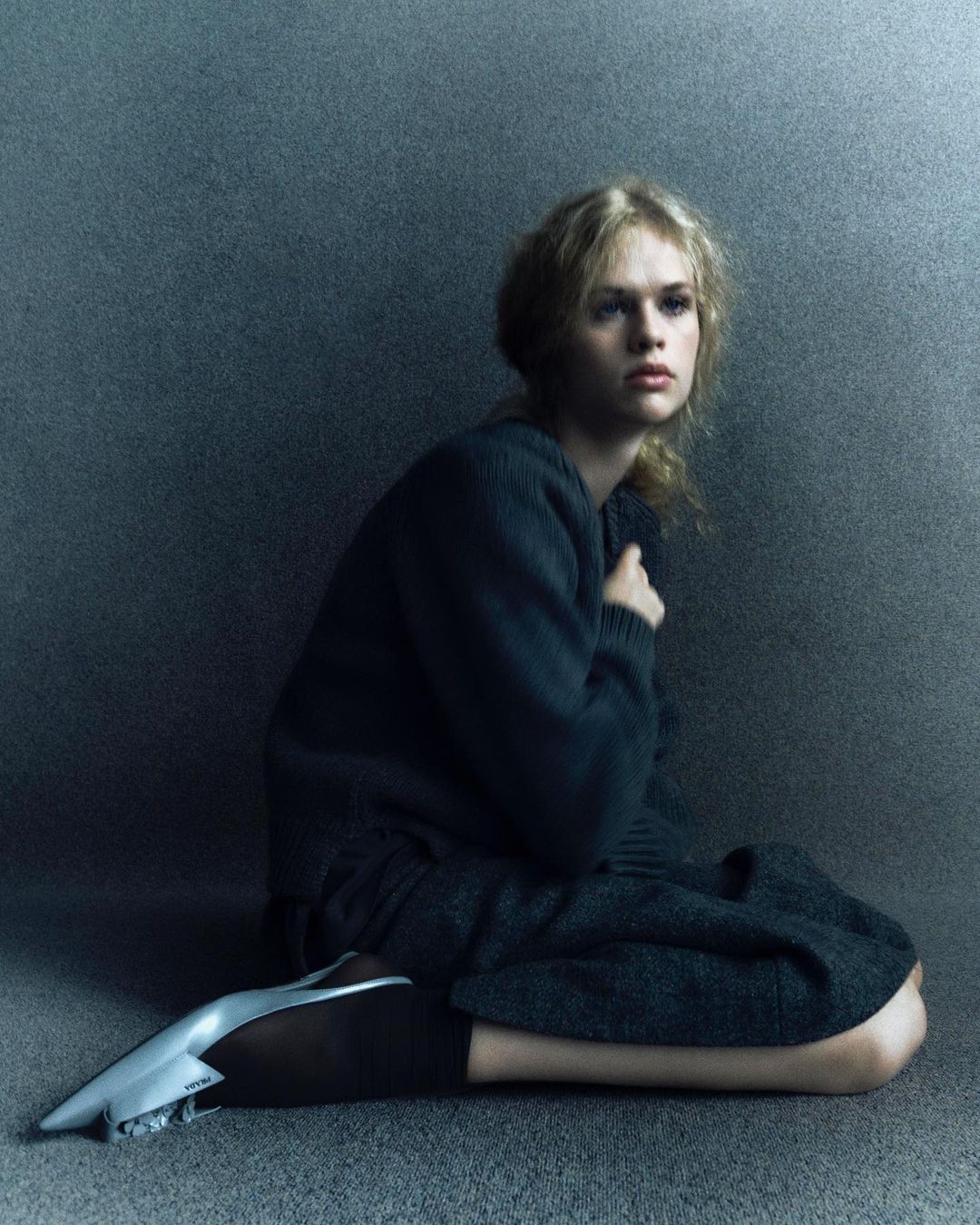 A woman wearing a dark sweater and skirt seated against a textured grey studio background.