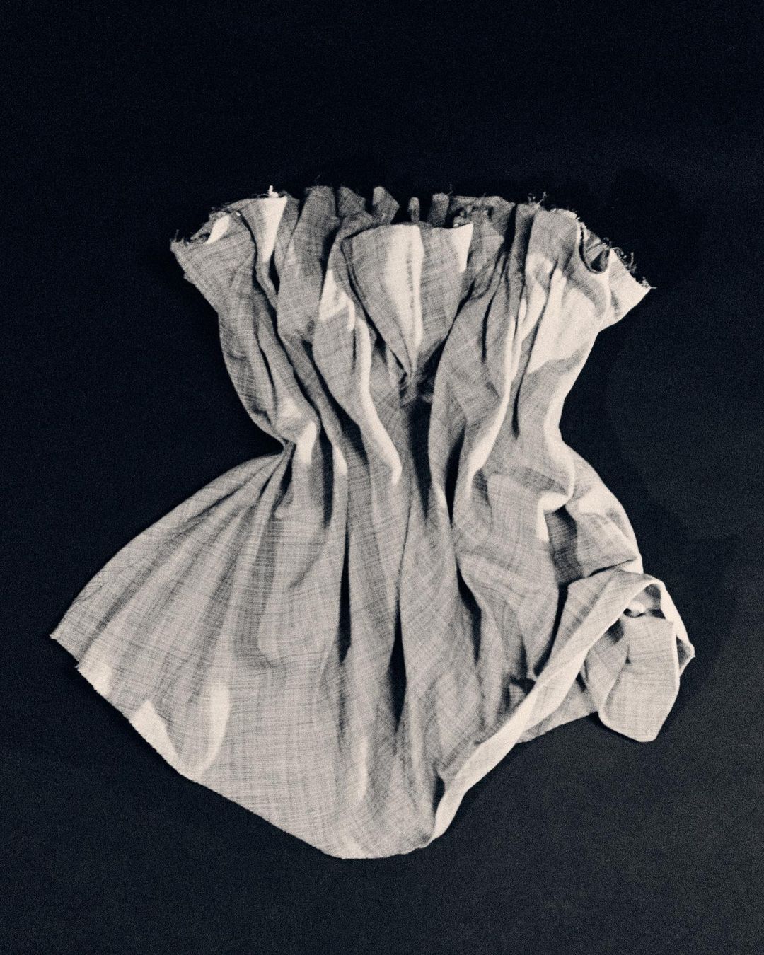 Textured grey and white fabric garment arranged in sculptural folds against a dark background by photographer Hanna Tveite.