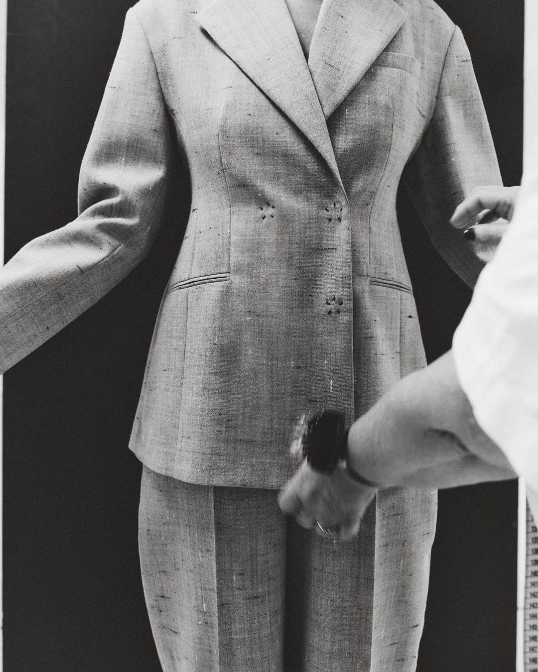 Tailored grey pant suit is closely examined by a hand wearing a dark watch, on a dark background with measuring tape on the right.