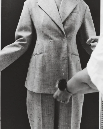 Tailored grey pant suit is closely examined by a hand wearing a dark watch, on a dark background with measuring tape on the right.