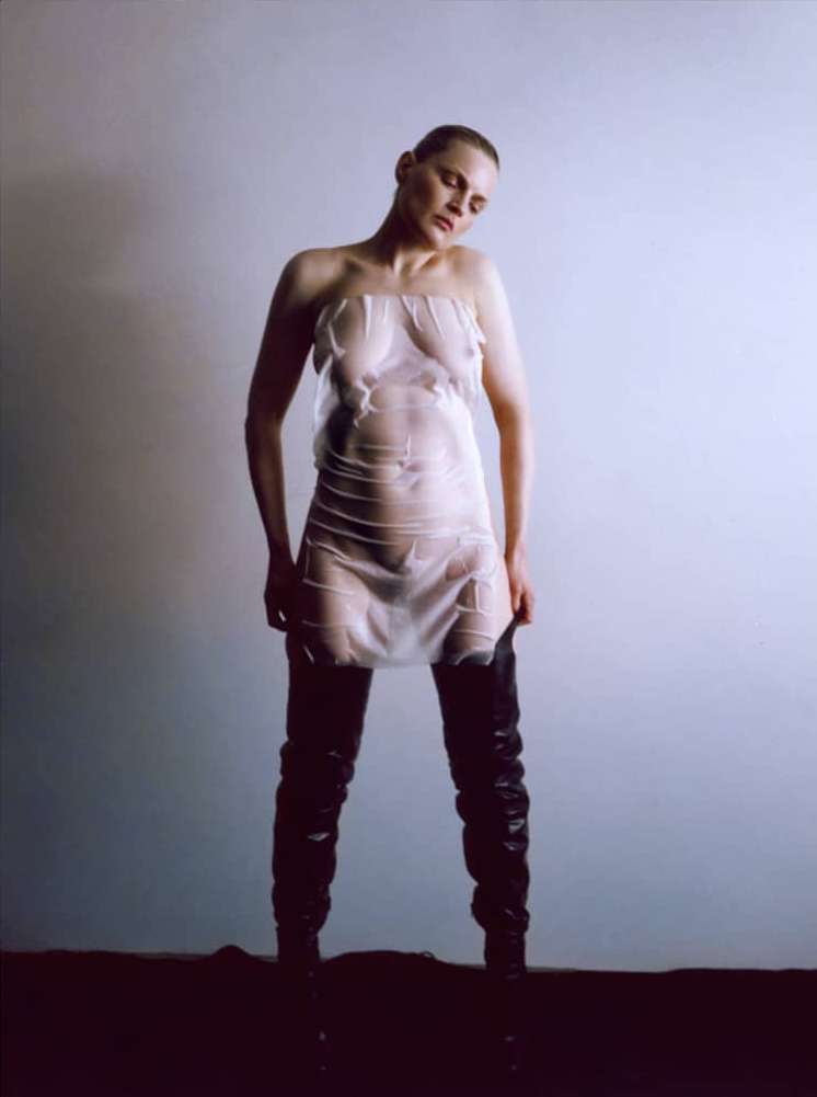 Model with shaved head wearing sheer plastic strapless mini dress and thigh-high black boots against plain background