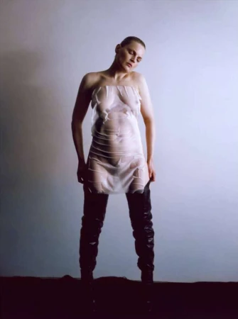 Model with shaved head wearing sheer plastic strapless mini dress and thigh-high black boots against plain background