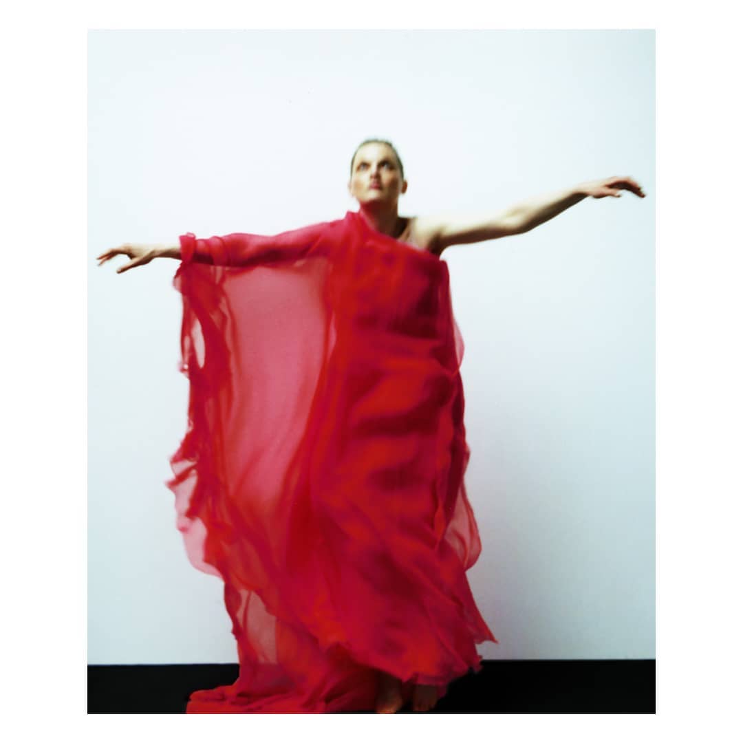 Elegant woman in red dress with arms outstretched in a studio setting. The full-length portrait captures movement and artistic style against a neutral background.