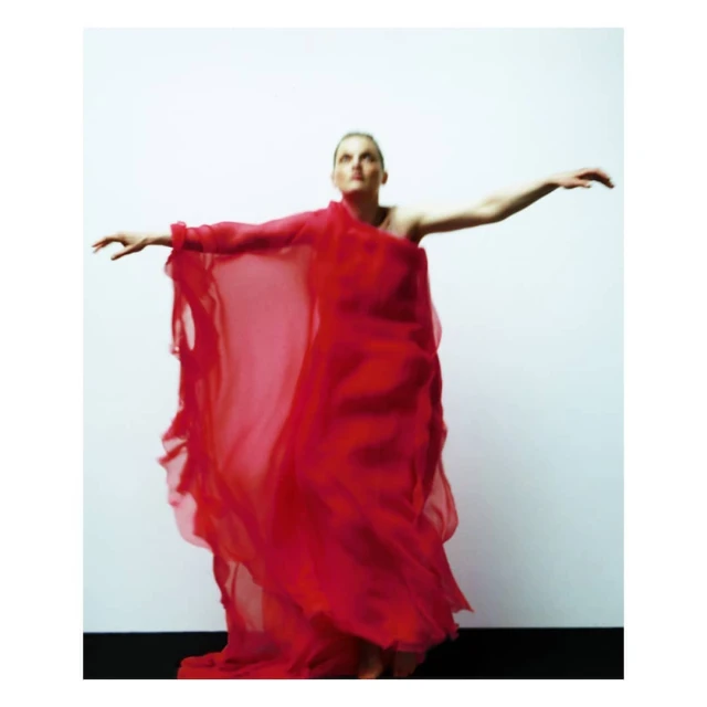Elegant woman in red dress with arms outstretched in a studio setting. The full-length portrait captures movement and artistic style against a neutral background.