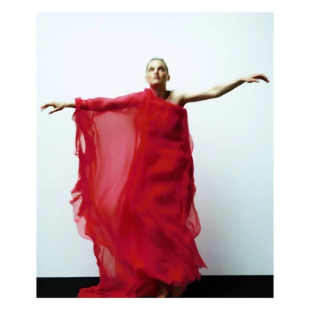 Elegant woman in red dress with arms outstretched in a studio setting. The full-length portrait captures movement and artistic style against a neutral background.