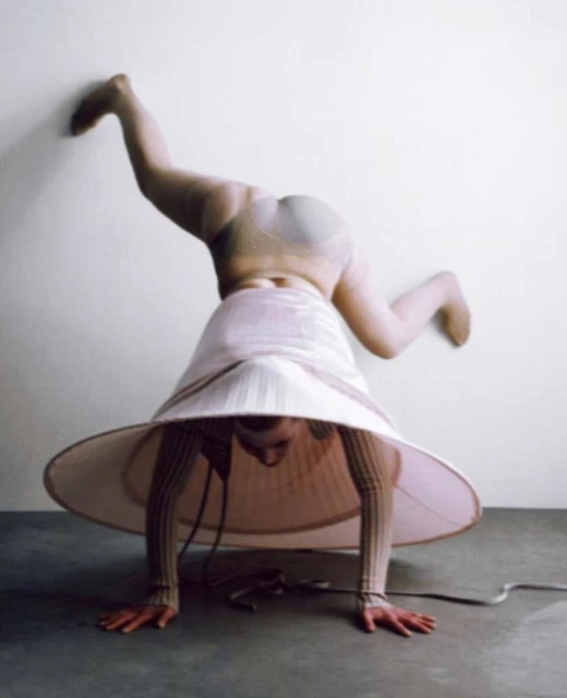 A woman in a lampshade skirt balances on her hands with her feet touching the wall, creating a visually striking and unusual composition.