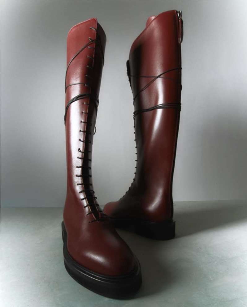 Pair of knee-high, lace-up maroon leather boots with a platform sole, set against a gradient grey background in a fashion product shot.