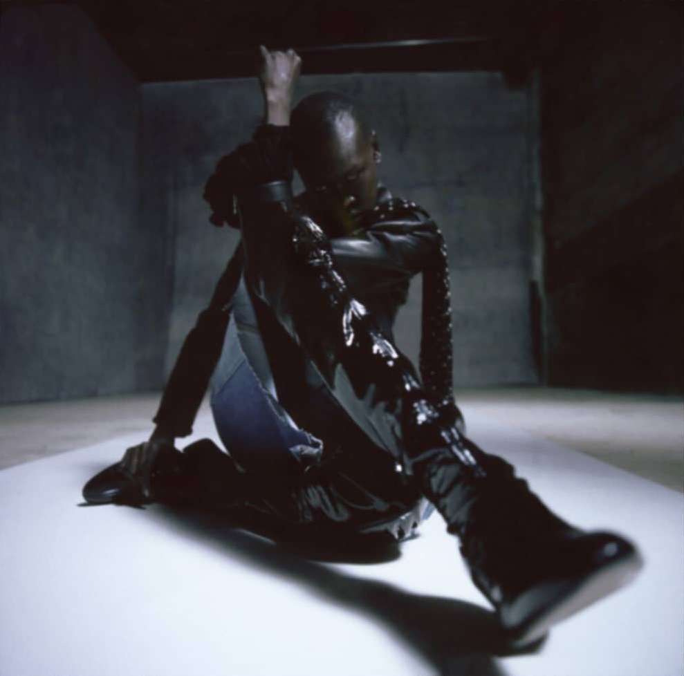 Adult male poses on a white surface in a dark room. He wears a black, glossy jacket and boots, casting a shadow in a dimly lit space.