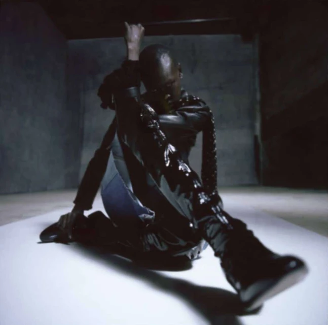 Adult male poses on a white surface in a dark room. He wears a black, glossy jacket and boots, casting a shadow in a dimly lit space.